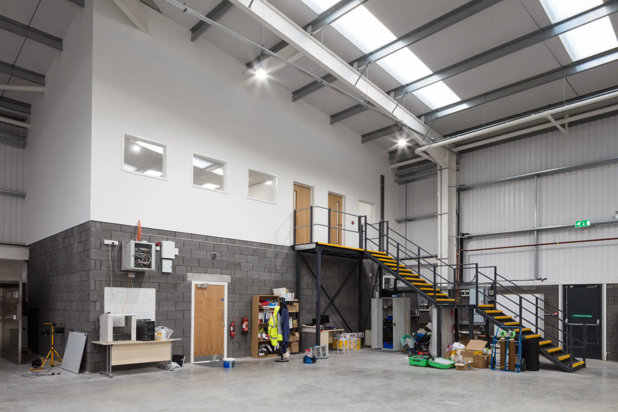 Modern warehouse interior with mezzanine office space, metal staircase, timber doors, internal office windows, industrial lighting, and polished concrete flooring.