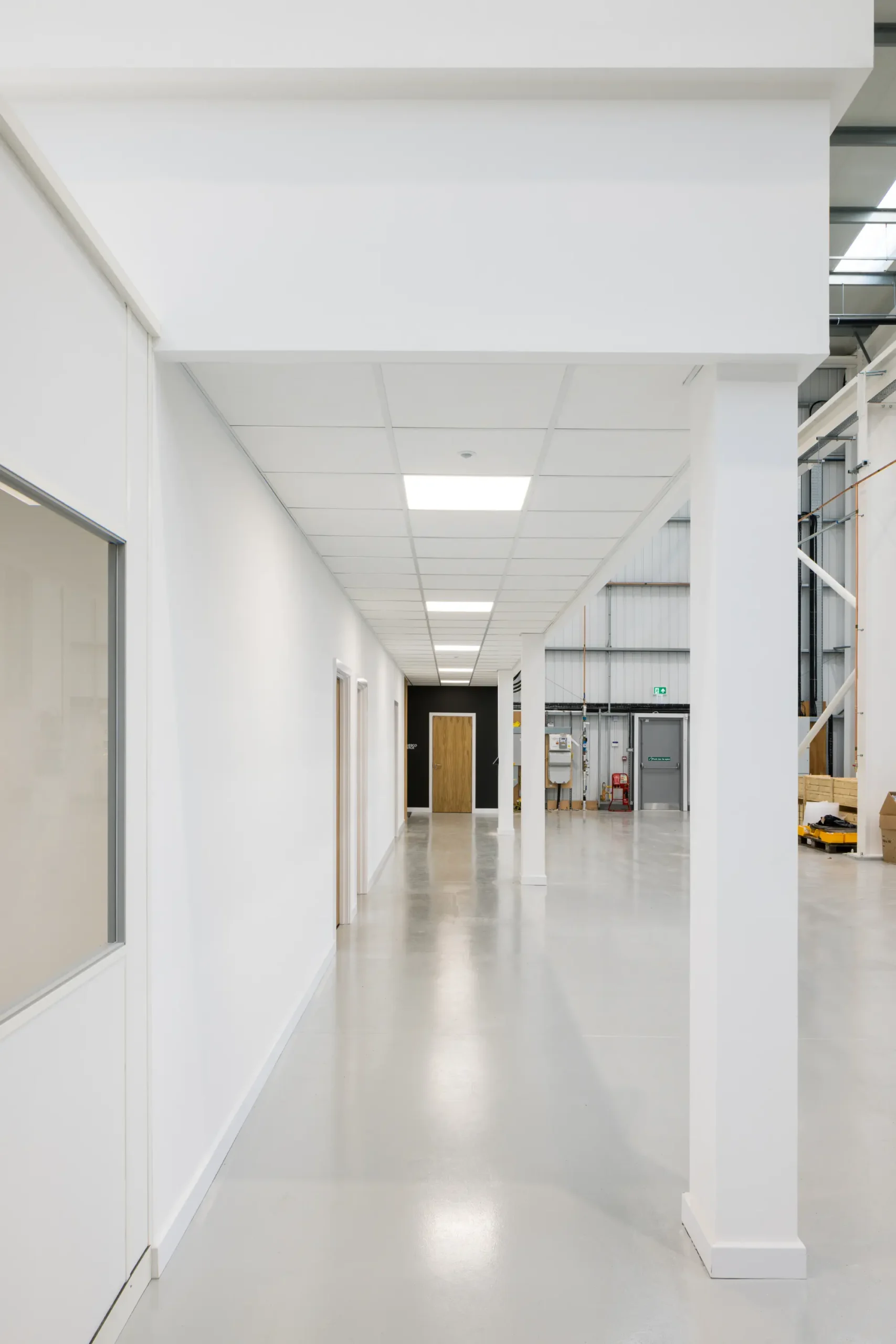 Long, clean corridor inside a modern industrial warehouse, with white walls, ceiling tiles, multiple doors on the left, structural columns, and an open warehouse space visible on the right