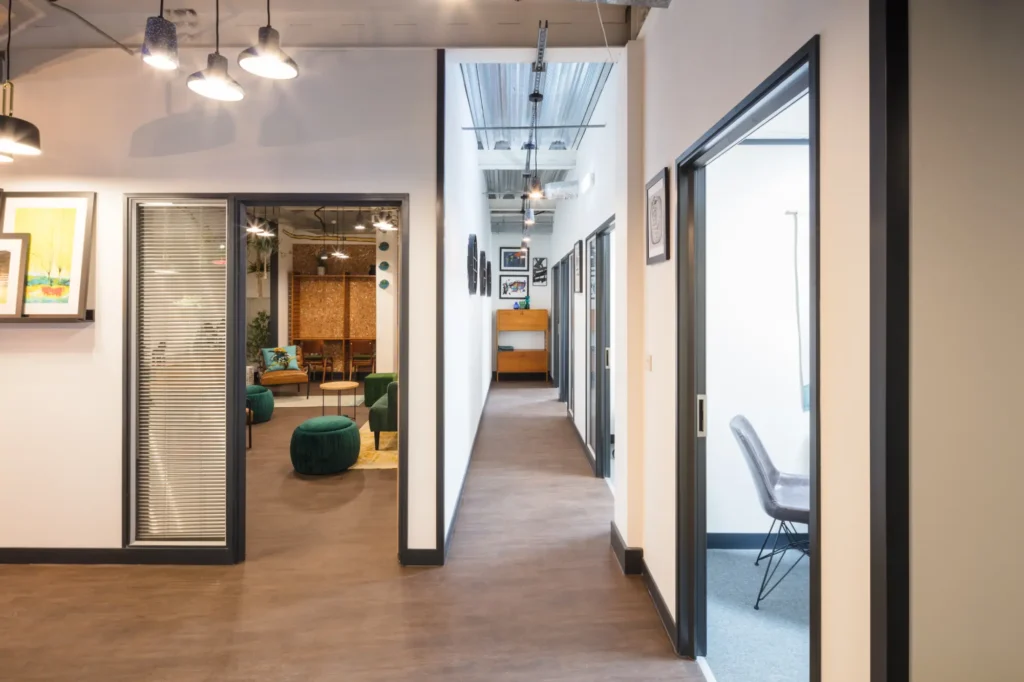 "Modern office space with multiple rooms, contemporary lighting, and wall art. View into a meeting room with table and chairs, featuring clean white walls and a polished floor."