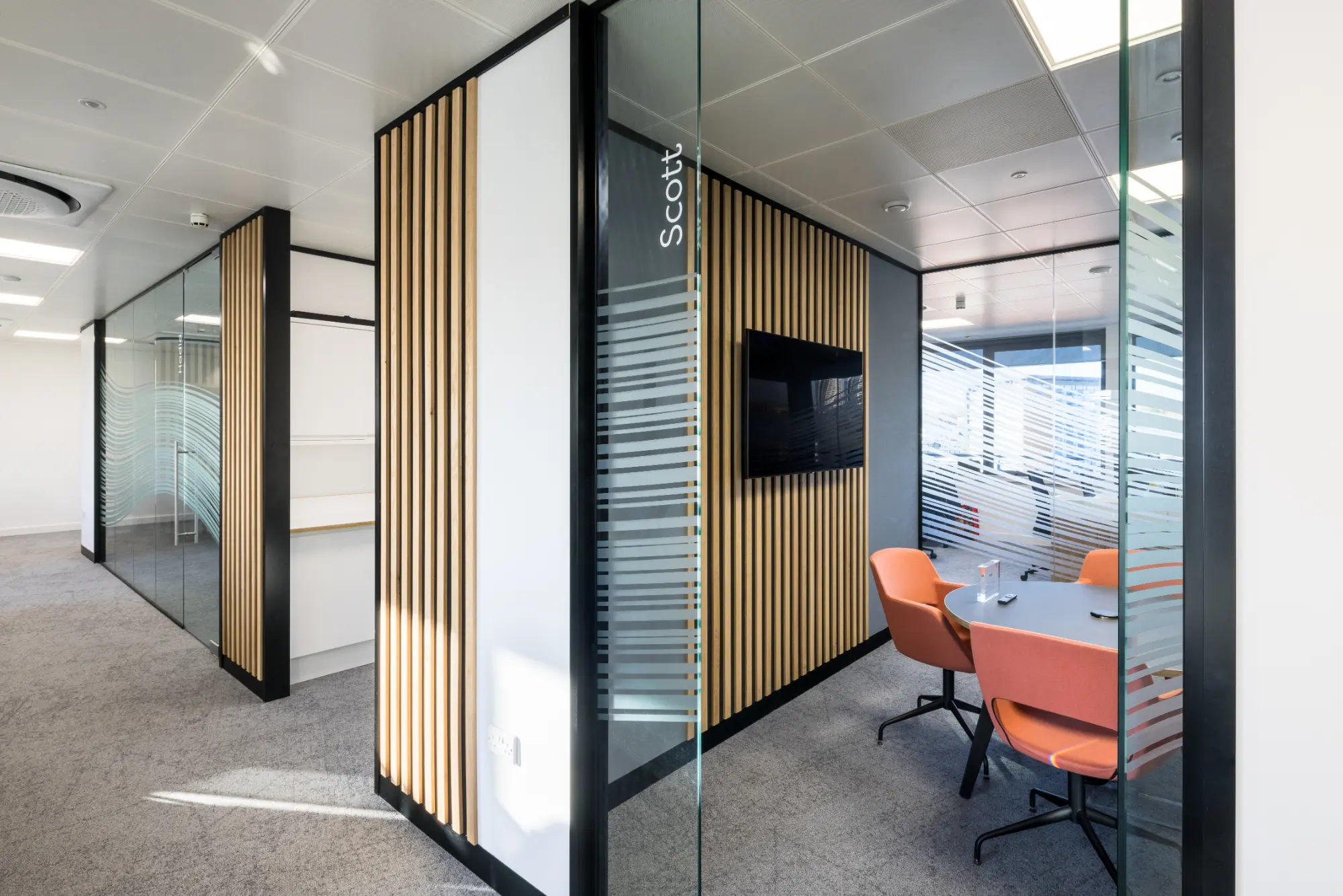 Two meeting rooms featuring wooden panels inside and out, each with a large TV, table, red chairs, and a visible room name.