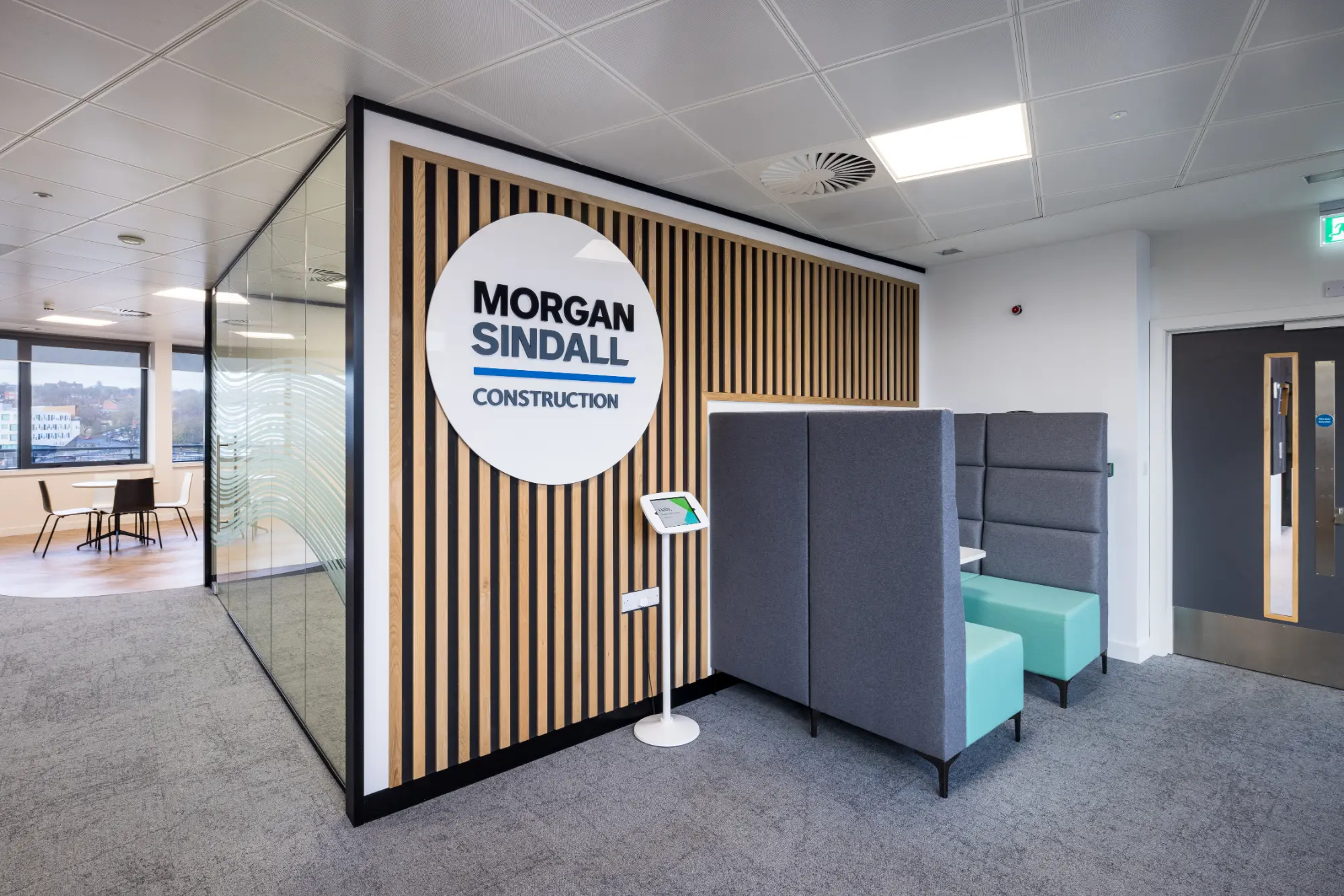 View of a meeting room from outside, showing a glass partition with a wooden panel wall displaying the Morgan Sindall logo, with chairs and table positioned outside the room.”