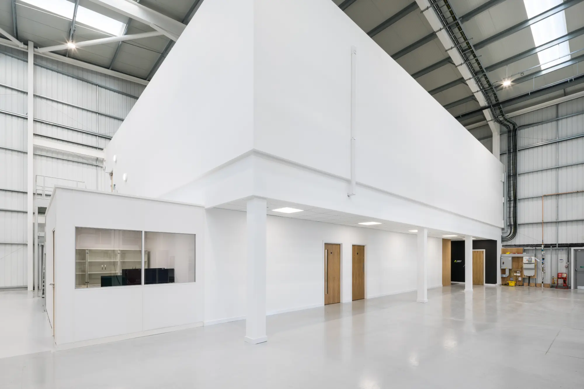 Spacious industrial warehouse interior with high ceilings, white walls, polished concrete floors, and a modern office pod with large windows and wooden doors.