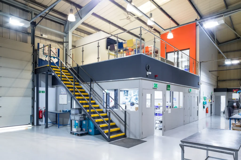 Interior of a modern industrial warehouse with steel-partitioned offices on the ground floor, a mezzanine office with glass railings, and a staircase with yellow safety markings.
